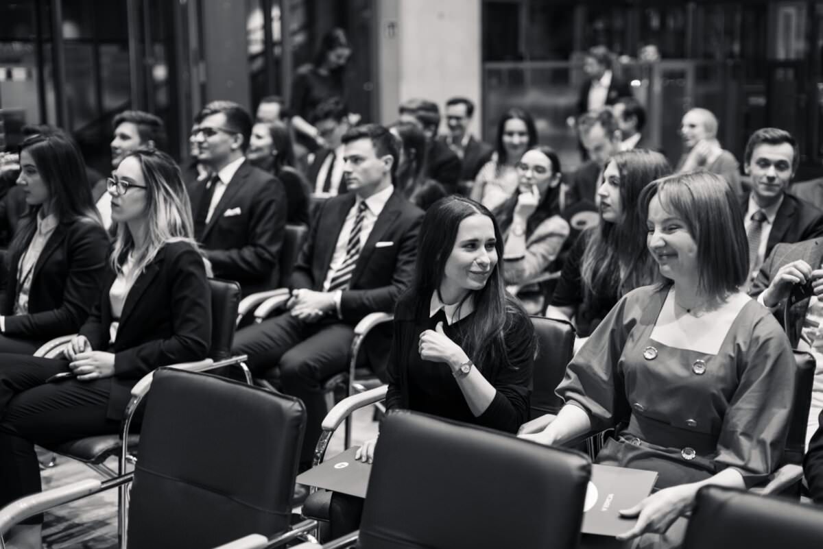 Schwarz-Weiß-Foto einer Gruppe von Menschen, die auf Stühlen sitzen, sich unterhalten und auf den Beginn der Veranstaltung warten. Die Szene wirkt förmlich, die Teilnehmer sind in Business-Kleidung gekleidet. Die Atmosphäre scheint lebhaft und voller Vorfreude zu sein, was die Essenz der Eventfotografie von Eventfotograf Warschau einfängt.
