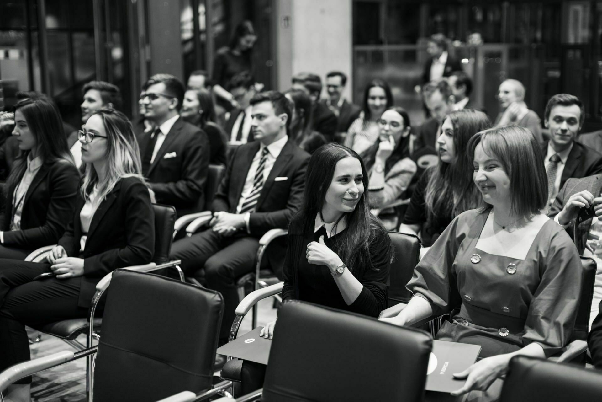 Black and white photo of a group of people sitting on chairs, talking and waiting for the event to begin. The scene appears formal, the participants dressed in business attire. The atmosphere seems lively and full of anticipation, capturing the essence of event photography by event photographer Warsaw.  