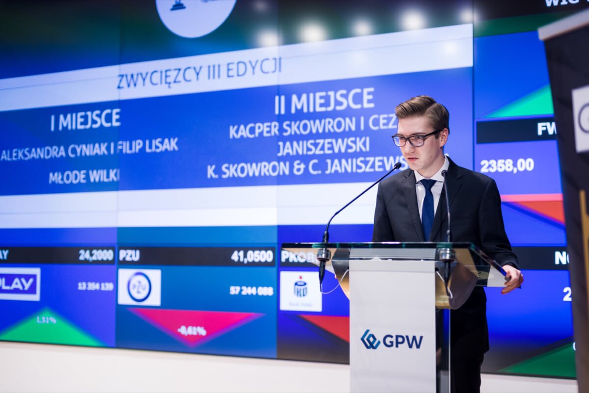 Ein junger Mann in einem Anzug steht an einem Podium mit einem Mikrofon und spricht. Hinter ihm befindet sich ein großer digitaler Bildschirm, auf dem die Namen und die Rangfolge der Ereignisse angezeigt werden, mit Text auf Polnisch. Auf dem Podium ist das WSE-Logo zu sehen. Die offizielle Veranstaltung könnte Teil einer Event-Fotoreportage eines Eventfotografen warszawa sein, die die Essenz der Veranstaltung einfängt.