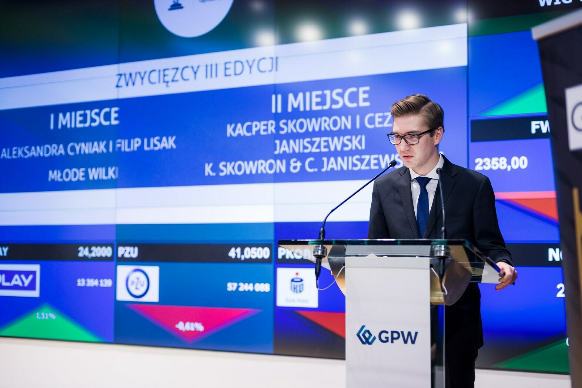 A young man in a suit stands at a podium with a microphone and speaks. Behind him is a large digital screen displaying the names and rankings of events, with text in Polish. On the podium is the WSE logo. The official event could be part of an event photo essay by an event photographer warszawa, which would capture the essence of the event.   