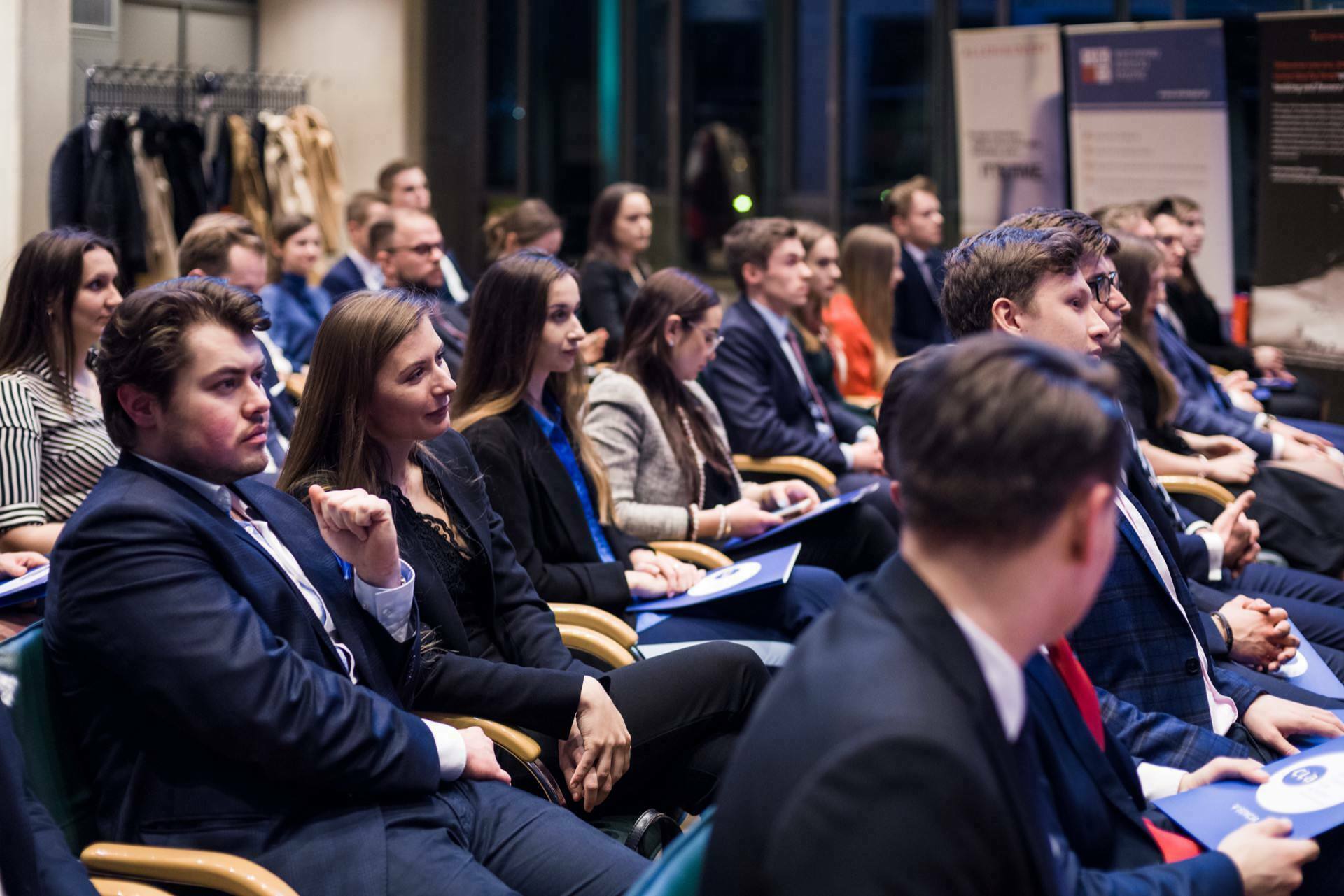 A group of men and women dressed in formal business attire sit in rows and listen attentively to a speaker at a conference or seminar. They hold booklets and notebooks, while coats and jackets hang on hangers in the background, capturing the essence of event photography. 