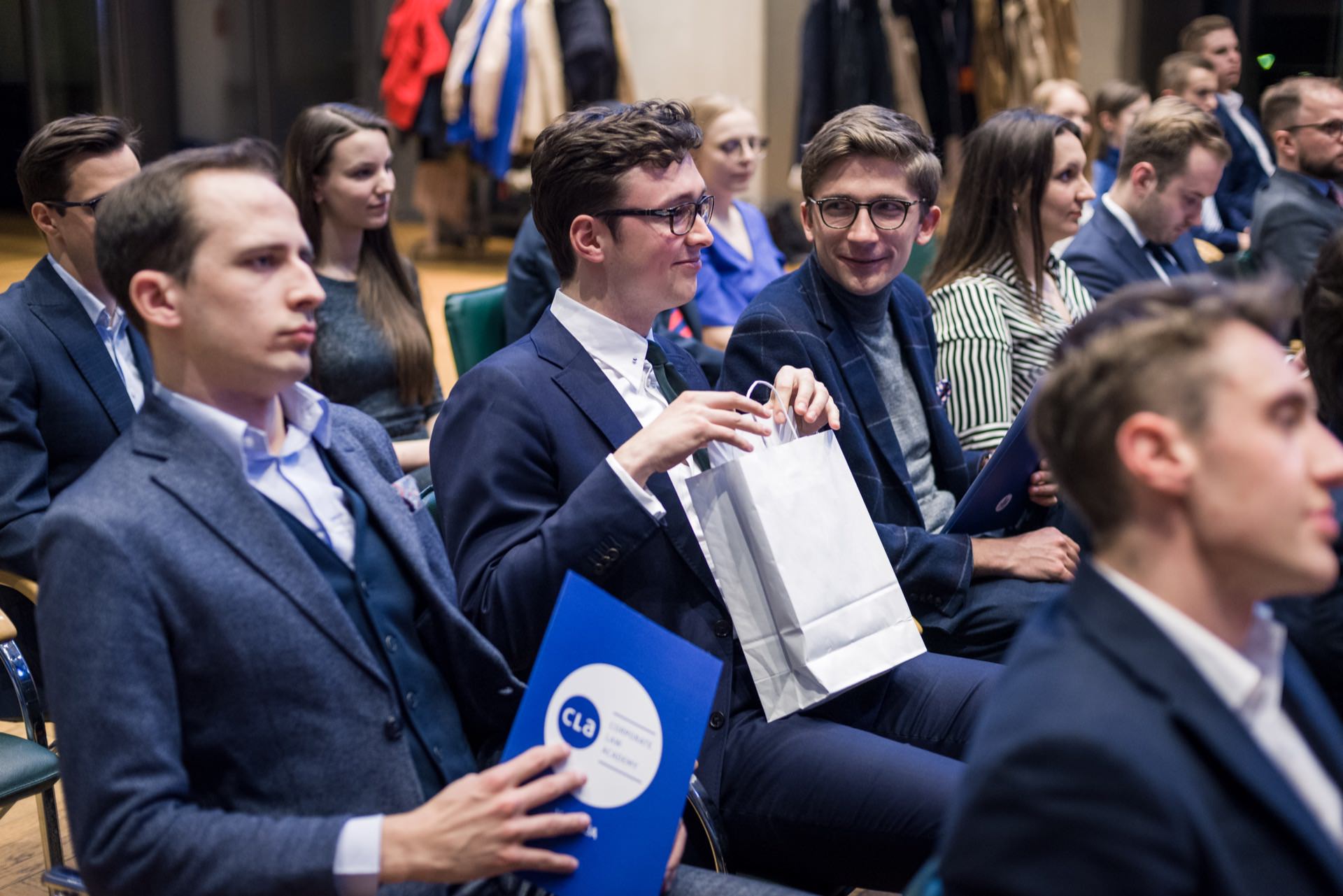 A group of professionally dressed adults sit in a conference room and look intently ahead. Some are holding blue briefcases, and one person in the middle is opening a white paper bag. Coats can be seen hanging in the background, capturing a moment perfect for photo opportunities.  