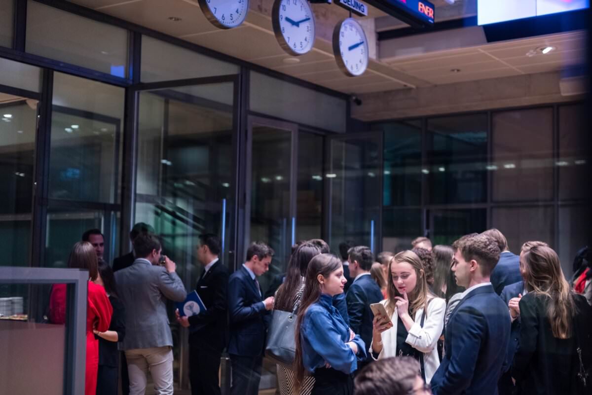 Eine Gruppe von Menschen in Geschäftskleidung unterhält sich in einem modernen Büro, perfekt eingefangen von einem Eventfotografen aus Warschau. Mehrere Teilnehmer versammeln sich in kleinen Gruppen, in eine Diskussion vertieft. Die großen Glasfenster des Raums und die Uhren, die die verschiedenen Zeitzonen anzeigen, tragen zur Dynamik der Eventfotografie bei.
