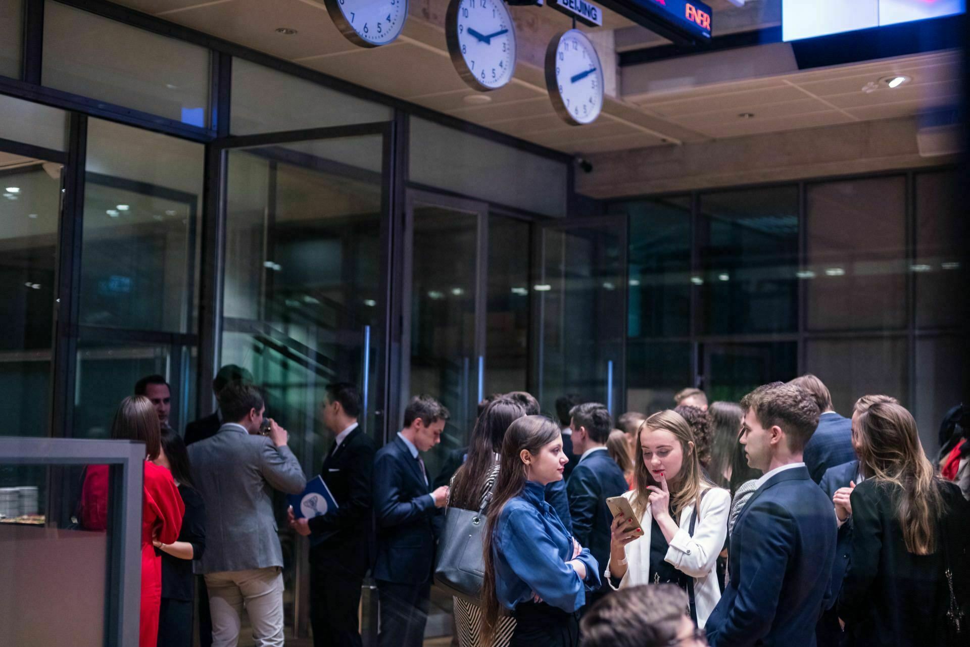 A group of people dressed in business attire are chatting in a modern office, as captured perfectly by an event photographer warszawa. Several participants gather in small groups, immersed in discussion. Large glass windows in the room and clocks showing different time zones add to the dynamic event photography.  