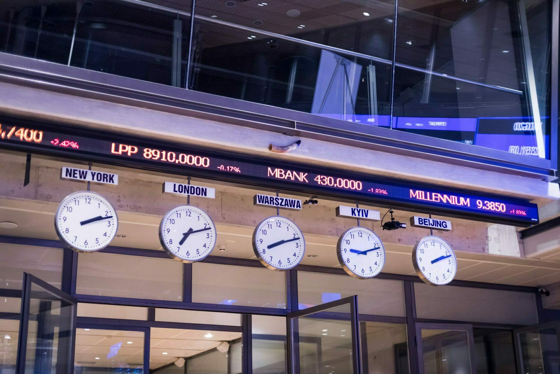 The stock market display shows various scrolling numbers and percentages in a modern building. Above, a number of clocks show the time in various cities such as New York, London, Warsaw, Kyiv and Beijing. The ceiling has a modern design with glass elements - perfect for event photography.  