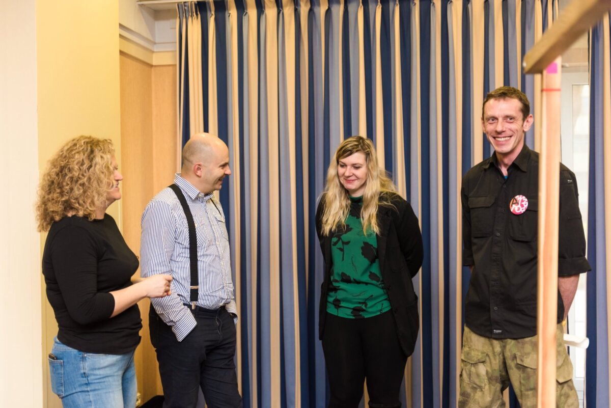 Four people are standing and talking in a room in front of a striped curtain. One woman on the left has curly hair and wears a black top. Two others, a man and a woman, stand in the middle, and on the right is another smiling man in a black shirt - a perfect spot for event photography.  