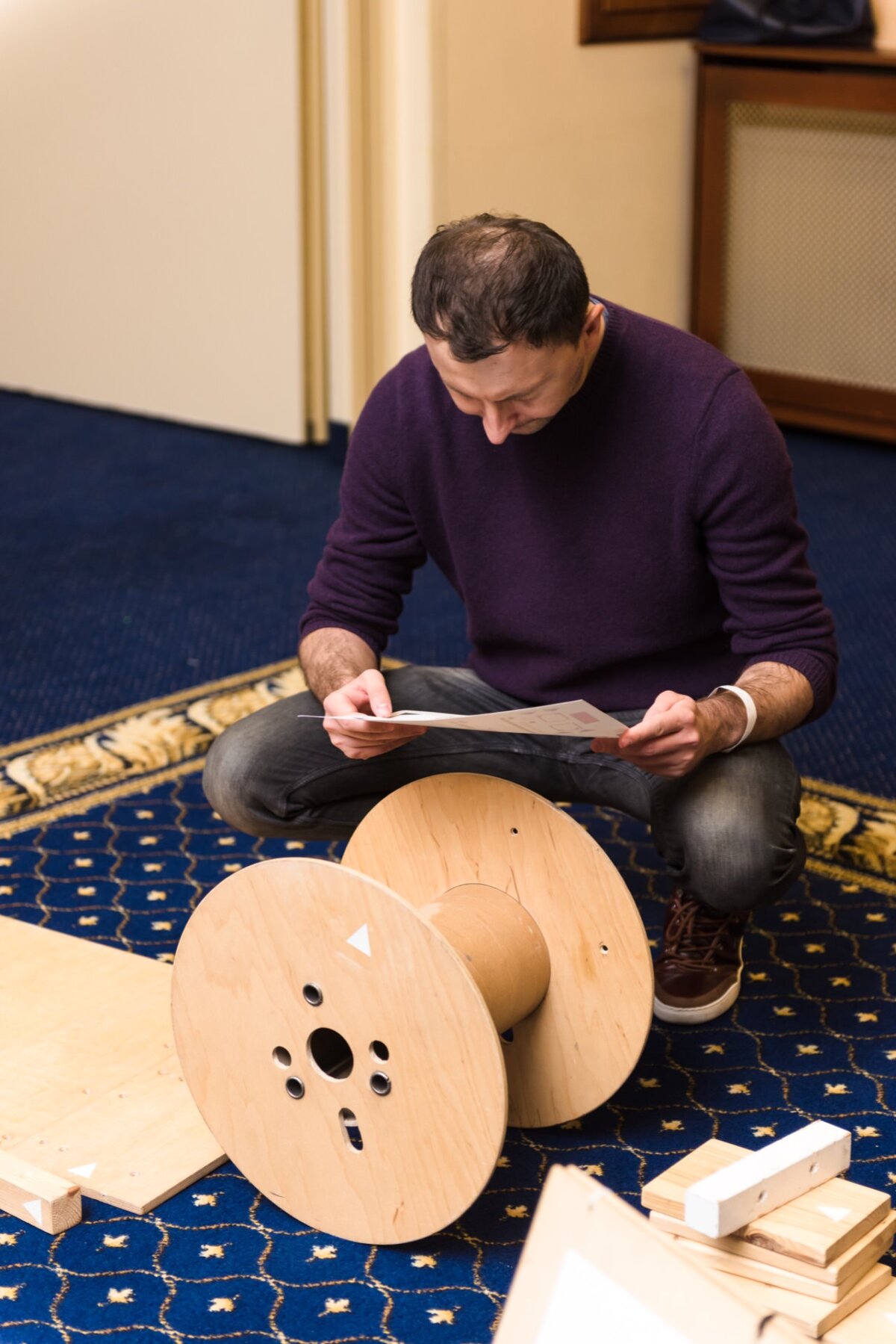 A man in a purple sweater and jeans crouches on a blue patterned carpet, carefully reading a piece of paper. Surrounding him are wooden items, including a large wooden spool, suggesting that perhaps he is assembling or inspecting something - perfect for photo coverage of events. 