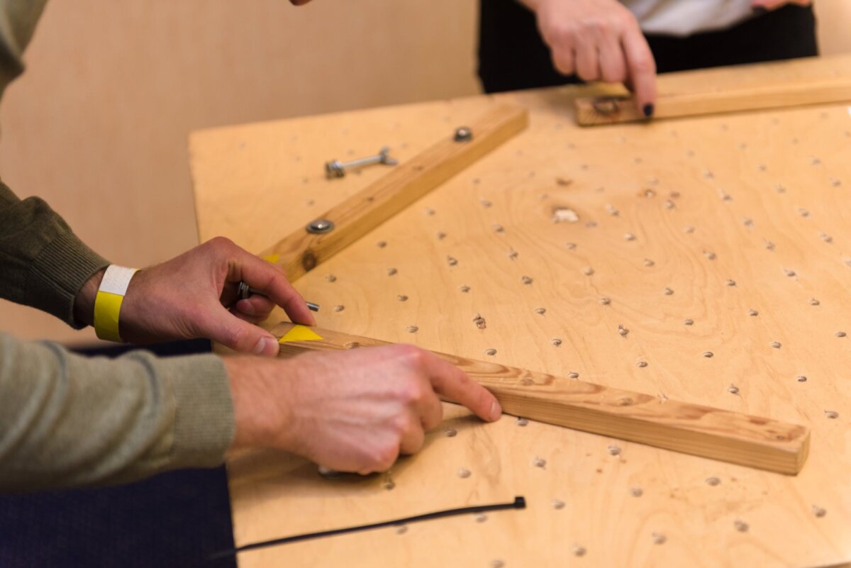 Two people are aligning and assembling wooden pieces on a perforated board. Hands in the foreground are adjusting a piece of wood, hands in the back are screwing in screws. The scene is ideal for photo coverage of events suggests an ongoing woodworking or DIY project.  