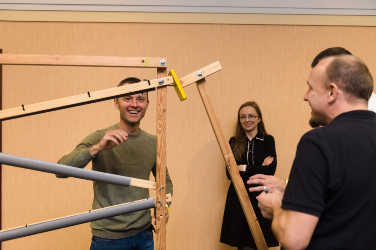 Four people interact with a wooden structure. A man in a green sweater and jeans is laughing and correcting it, capturing the spirit of the photo-report of events. Another person in a black shirt and jeans is turned face down, while a woman in a black dress and glasses smiles in the background.  