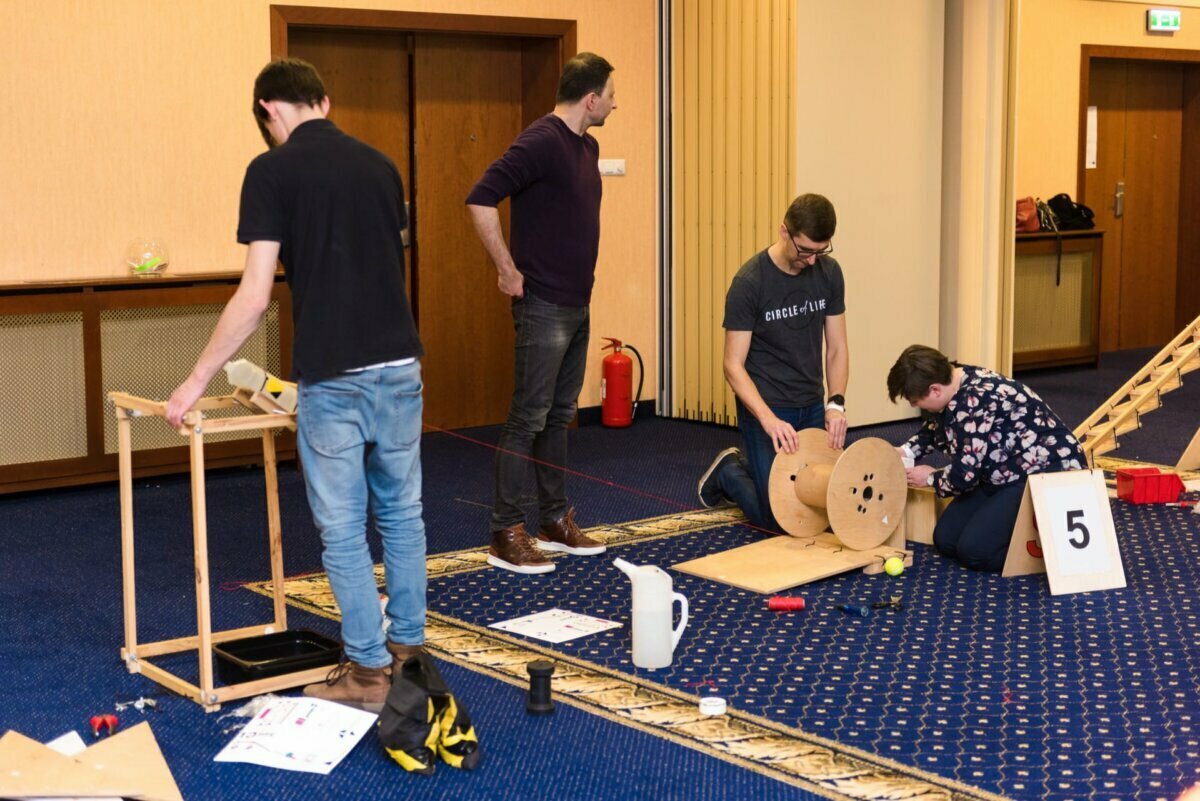 Four people are working to build a project using various materials, such as wooden spools and frames. One person stands and watches while three others assemble the objects on the ground. In the background is a beige wall, a door and a fire extinguisher. A "5" sign hangs on the floor - perfect for photo opportunities of the events.   