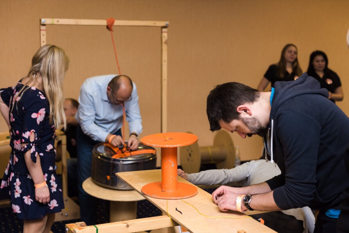 Adults engage in team building or workshop activities in the room. Participants perform a variety of tasks, including manipulating cords and handles connected to equipment standing on tables. A woman in a flowery dress watches as others focus on their tasks, creating the perfect scene for event photography.   