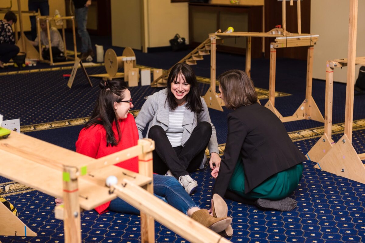 The three women are sitting on the carpet, having a lively conversation and enjoying each other's company. They are surrounded by wooden structures, presumably part of a Rube Goldberg machine - a perfect moment captured for a photo essay of the event. One woman is wearing a red sweater, another a gray sweater and the third a black jacket.  