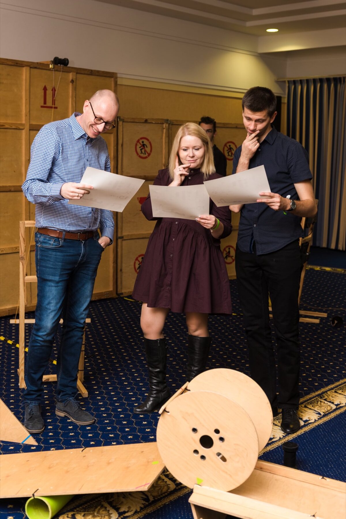 Three people are standing in a room with wooden boxes and unfinished wooden projects. They are holding papers and seem to be discussing something. The scene captured by the event photographer warszawa shows them deeply involved and thoughtful.  