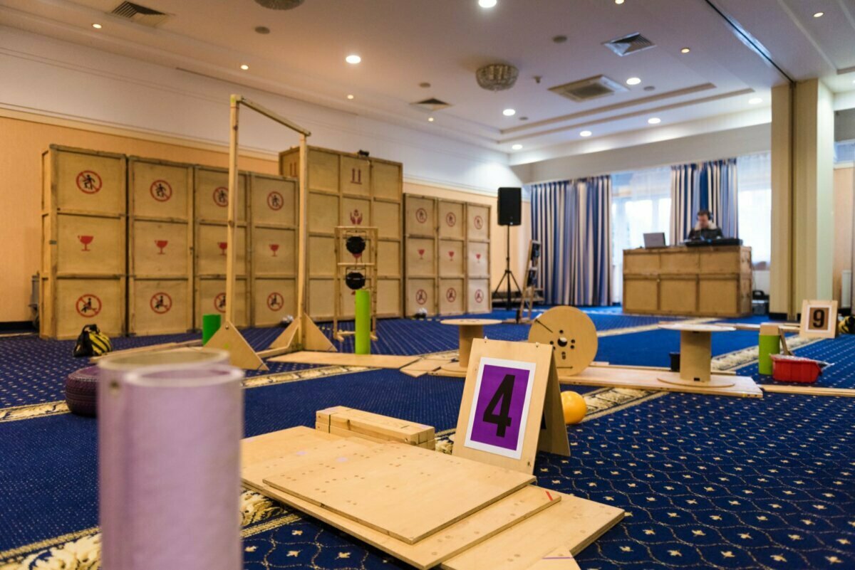 A brightly lit room with a carpeted floor, equipped with a miniature golf course or obstacle course. Various wooden structures and challenges are placed throughout the facility, numbered with signs, including the prominently placed purple number 4. In the background, a person is working at a table - ideal for photo coverage of events.  