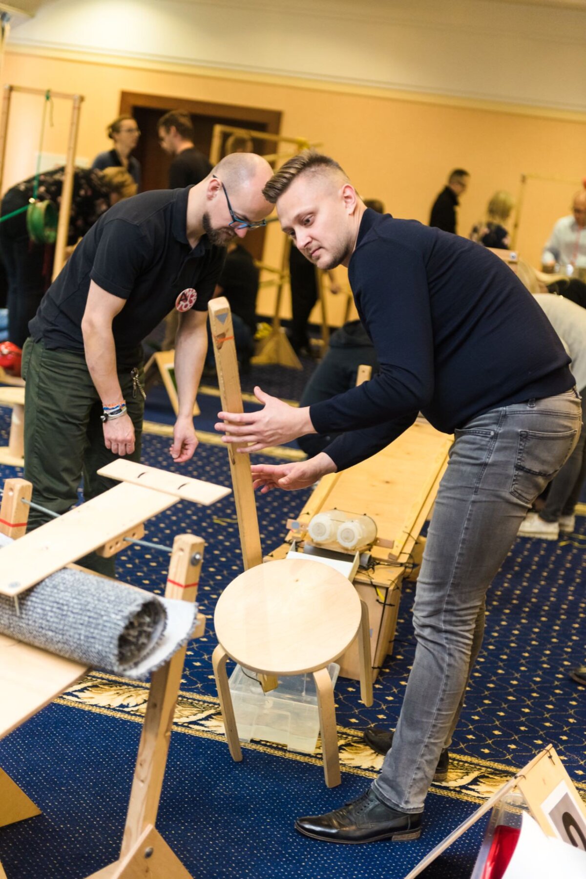 Two men are in a room constructing wooden objects. One, with a beard and glasses, is wearing a black t-shirt and green pants; the other, with short hair, is dressed in a black long-sleeved shirt and gray jeans. This scene could easily have been part of an engaging photo essay of events showcasing their craftsmanship among a few partially assembled objects and tools.  