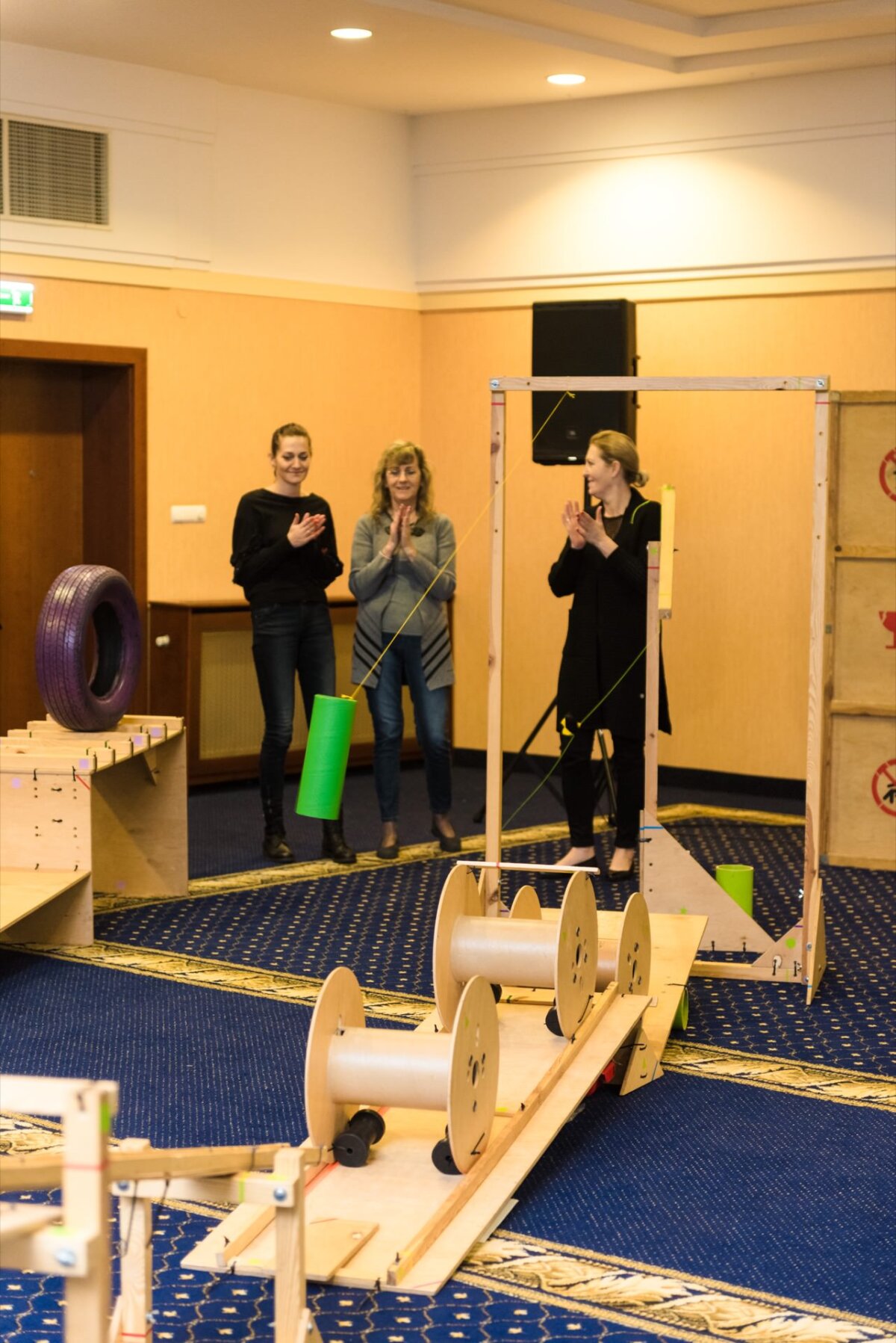 Three women stand in a room with light-colored walls and blue carpet, watching and clapping near a wooden structure equipped with spools and ramps. One woman holds a green cylindrical object. The installation looks like part of an interactive exhibition or event, captured in this immersive photo essay of events.  