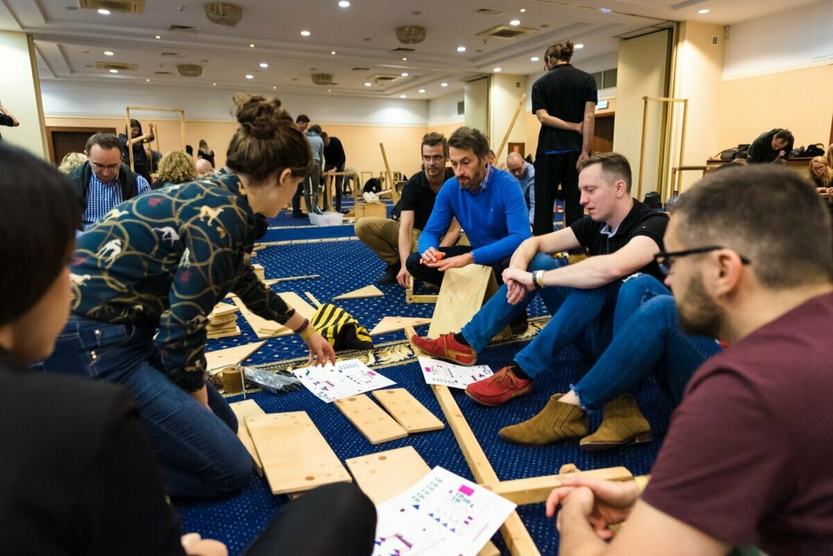 A group of people sit on the floor of a conference room and undertake a joint activity. Surrounded by wooden boards, paper instructions and tools, everyone seems focused on the task at hand. The group's attention is directed by one woman. This scene captures a perfect photo-op of the events.   