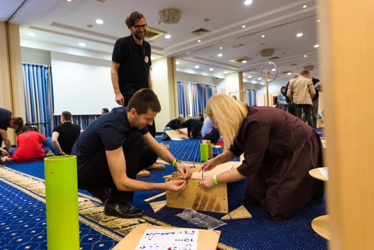 In this photo report of events, two people are assembling wooden pieces on the conference room floor, while other participants can be seen in the background. The person on the left kneels and sets up the block, the person on the right secures it. Both are focused on the task at hand.  