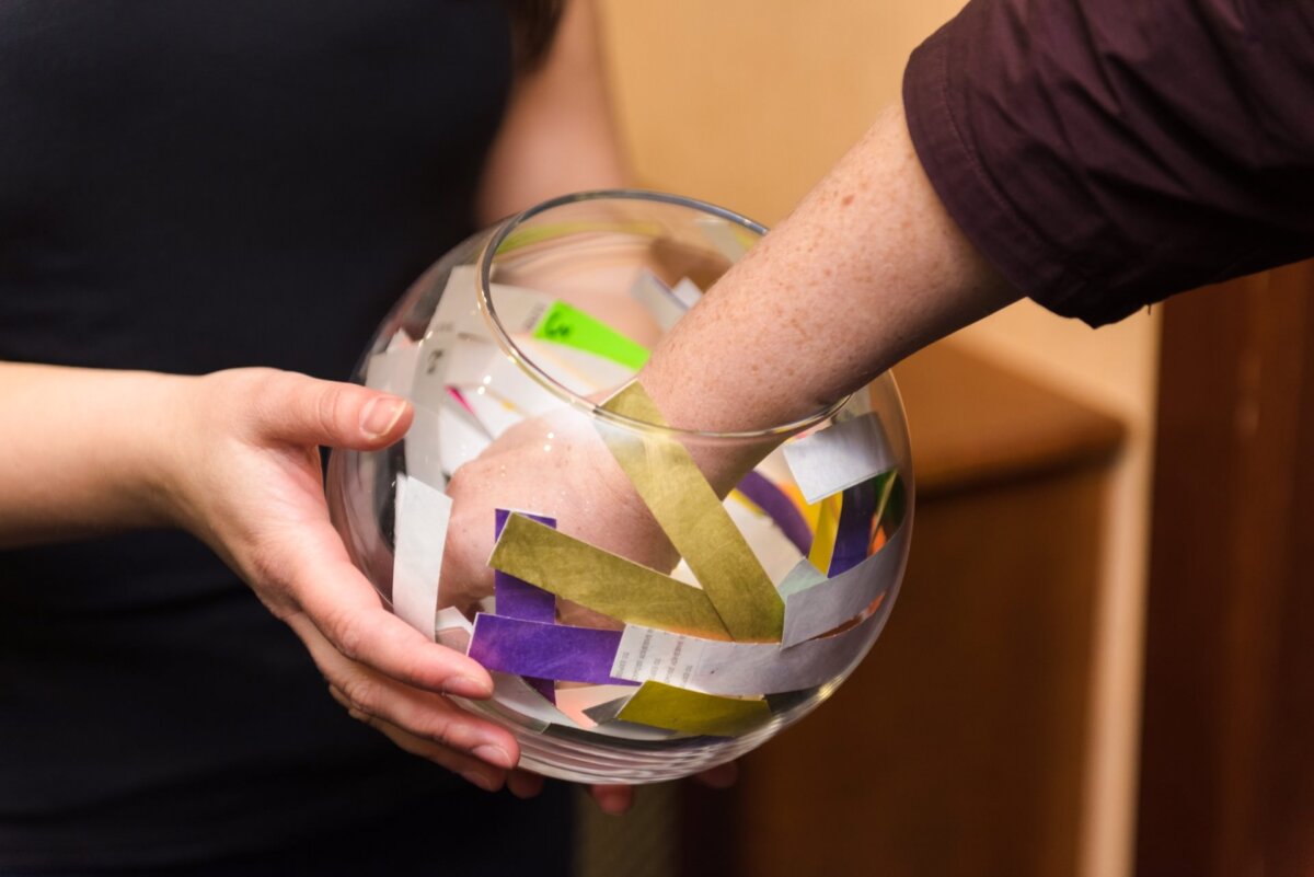 One person's hand reaches into a glass bowl filled with colored paper strips while another person holds the bowl. The setting appears to be indoors. The transparent bowl, captured by an event photographer Warsaw, reveals the vibrant paper strips.  