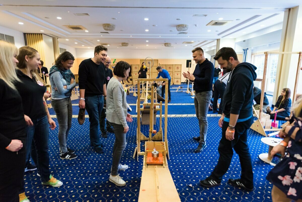 A group of people gather in a room and take part in an integration activity using a wooden device. They smile and interact with each other, suggesting teamwork and cooperation. The room has a blue carpet and large windows that let in natural light - perfect for event photography.  