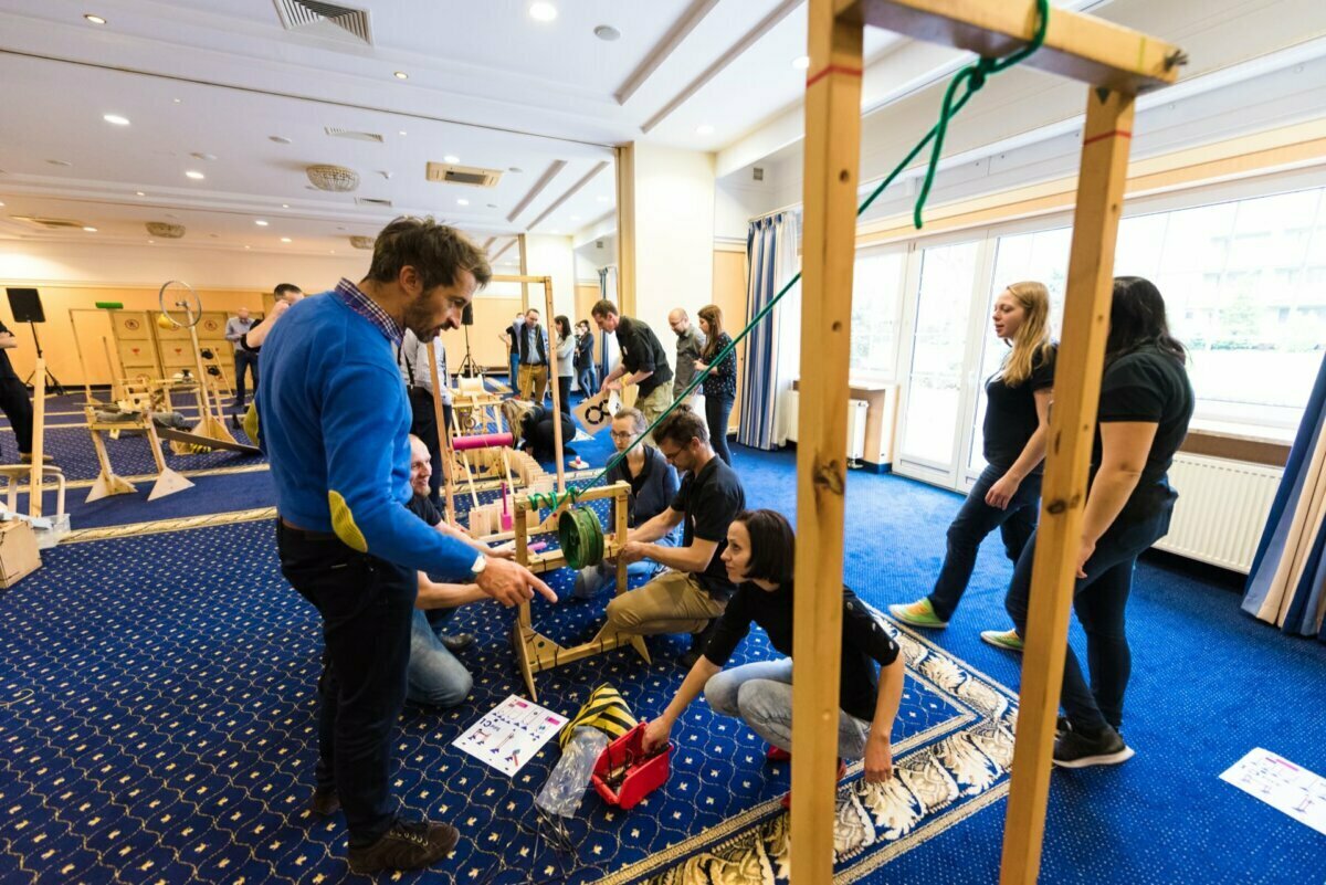 A group of people engage in hands-on team-building activities in a spacious, well-lit room. They work on wooden structures using ropes and other tools. Some participants kneel, some stand, and the instructor seems to guide them, which is ideal for event photography.  