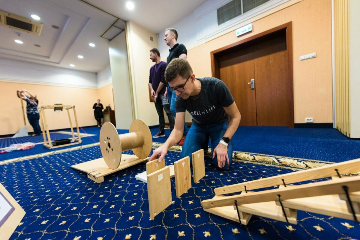 A man kneels on a blue carpet, arranging wooden pieces as part of a constructive project. Nearby, another person stands next to a large wooden spool. Two more people stand in the background against a beige wall and watch what's going on - a perfect scene for event photography that captures every last detail.  