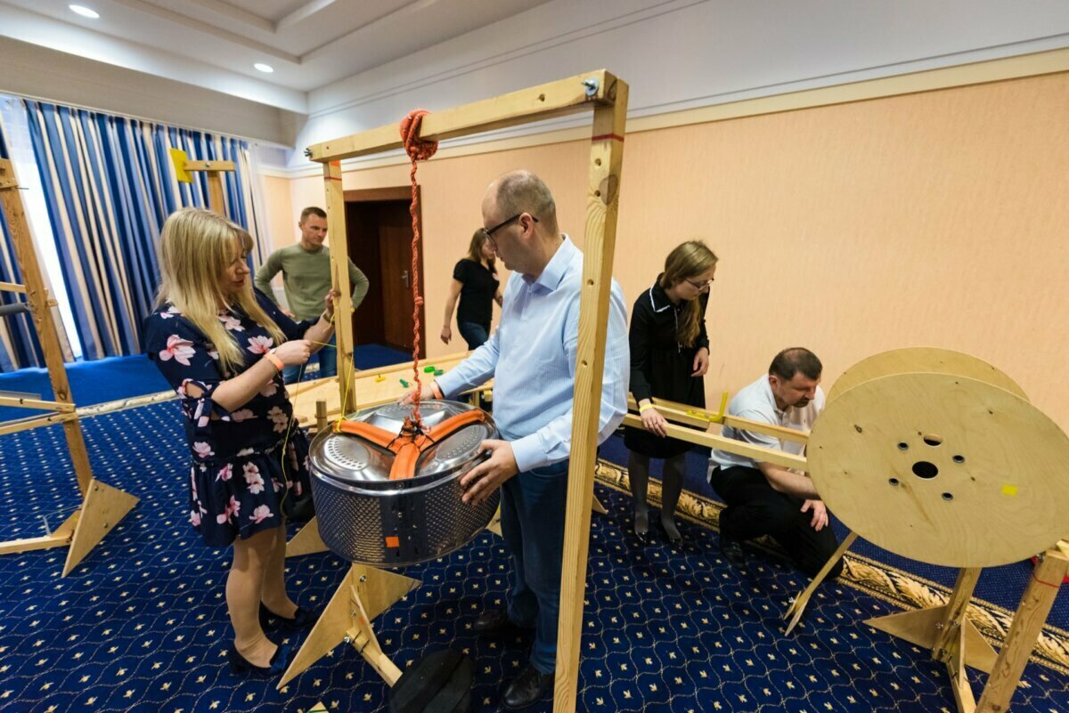 A group of people take part in a team-building activity in a room that uses ropes and wooden structures. They intensely work on lifting and maneuvering a large, round metal drum attached to a pulley system. The room, ideal for event photography, features a blue patterned carpet and striped curtains.  