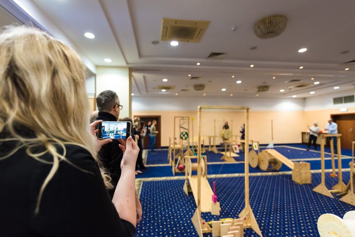 The person holding the phone is recording a miniature obstacle course for robots or models in a well-lit room with a blue carpet. The course features various wooden structures and ramps, and several people watch the setup in the background, creating an immersive photo essay of the events. 