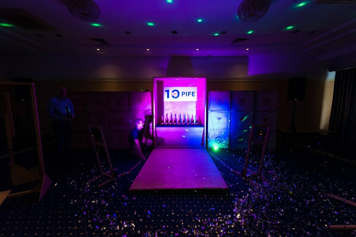 A dimly lit room with a wooden game set illuminated by purple and blue lights. The game features several bottles lined up in a row against a background with a "10 PIFE" sign. Confetti is scattered on the floor, and a person can be seen partially kneeling near the set - a perfect scene for event photography.  