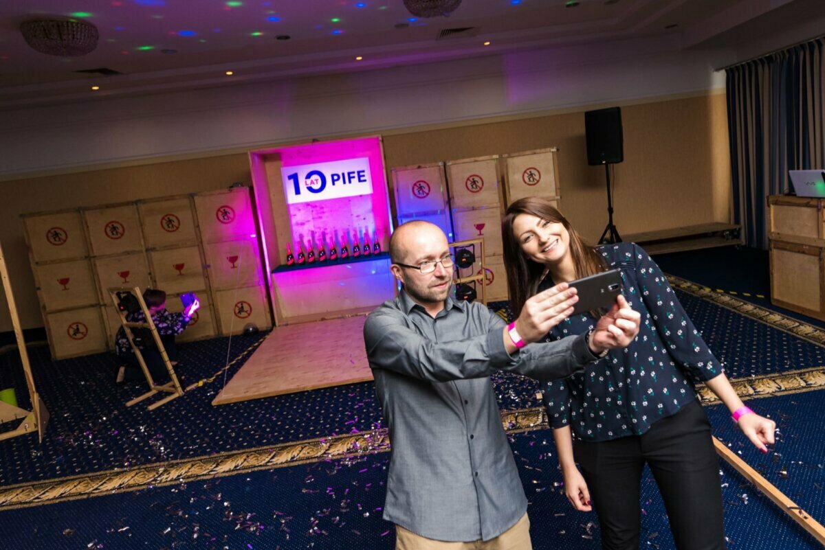 Two people, a bald man with glasses and a woman with long brown hair, take a selfie during an event taking place in a room. Behind them, the screen shows "10 PIFE" surrounded by colorful lights and heart-shaped decorations on the wall, and the floor is covered with confetti - a perfect scene for event photography in action. 