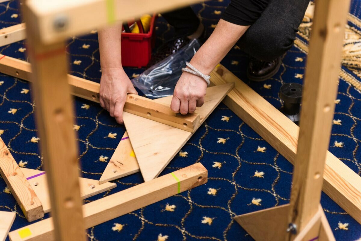 A person assembles wooden pieces of a triangular design on a patterned blue carpet. Numerous wooden beams and a small red toolbox with additional utensils are visible - perfect moments for photo-reportage of events. 