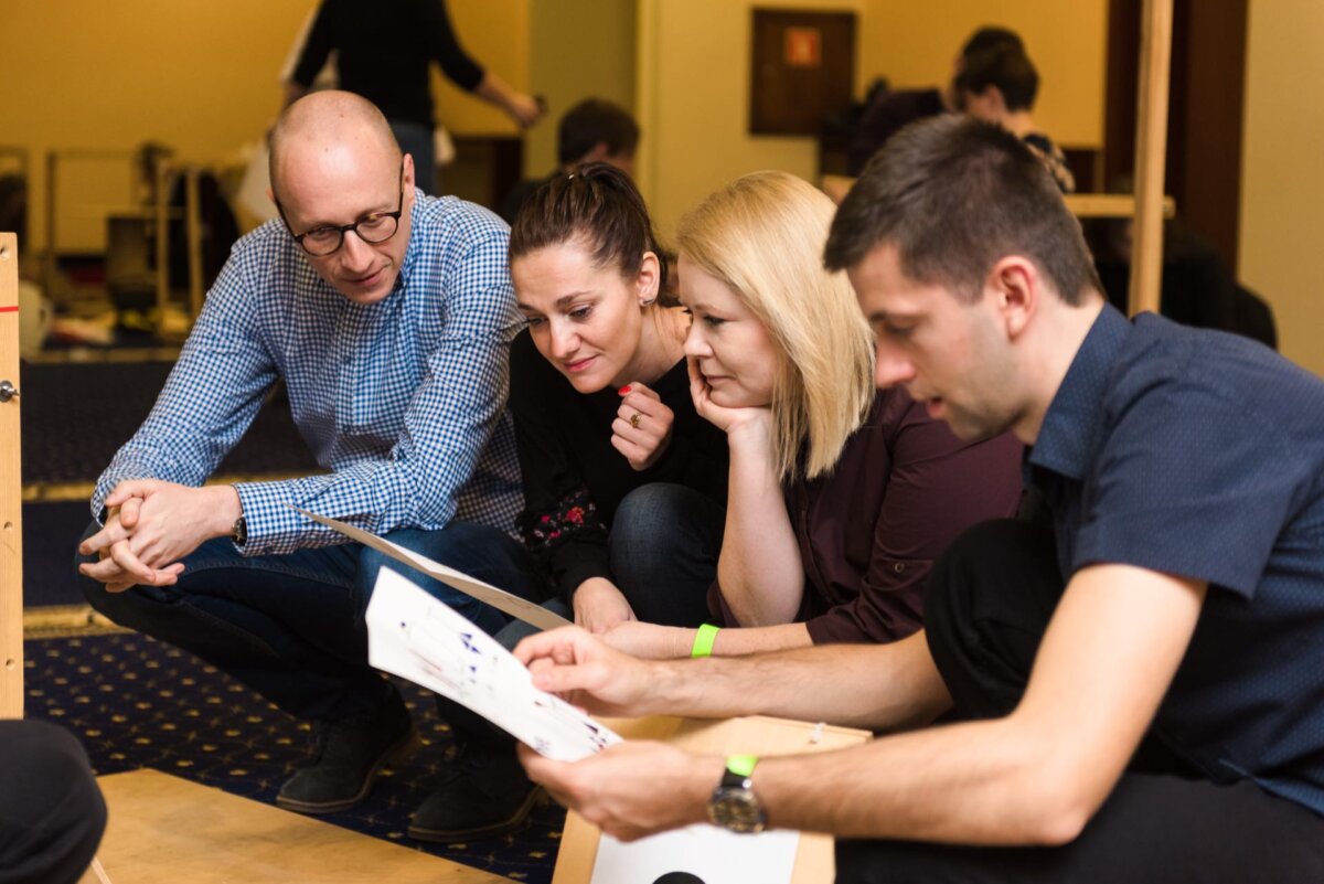 Four adults have gathered in a room, crowded around a piece of paper. One man kneels and holds the piece of paper, while the others sit or crouch, looking at it carefully. They look as if they are engaged in a discussion. Several more people can be seen in the background; it's the perfect moment for event photography.   