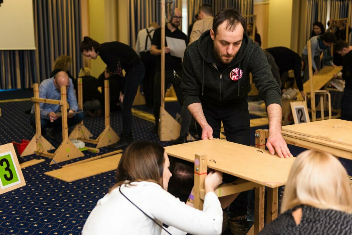 A group of adults engaged in hands-on team-building activities. They assemble wooden structures on a carpeted floor in a conference room. Key participants focus on their tasks, using tools and wooden panels. Some of the structures have visible numbers, ideal for event photo documentation.   