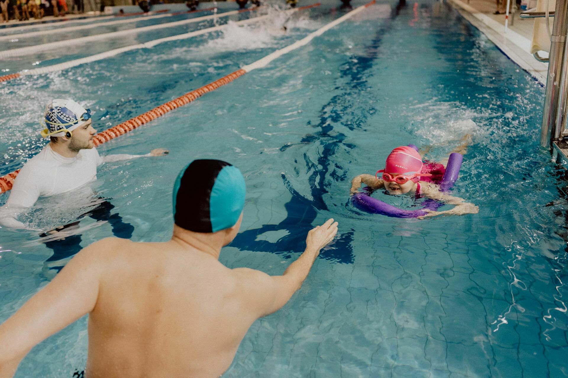 Two swimming instructors help a young child wearing a pink cap and swimming goggles in a pool. The child supports himself with a noodle, swimming toward one of the instructors, who reaches out for help. The event was captured perfectly, providing excellent photo coverage of the events.  