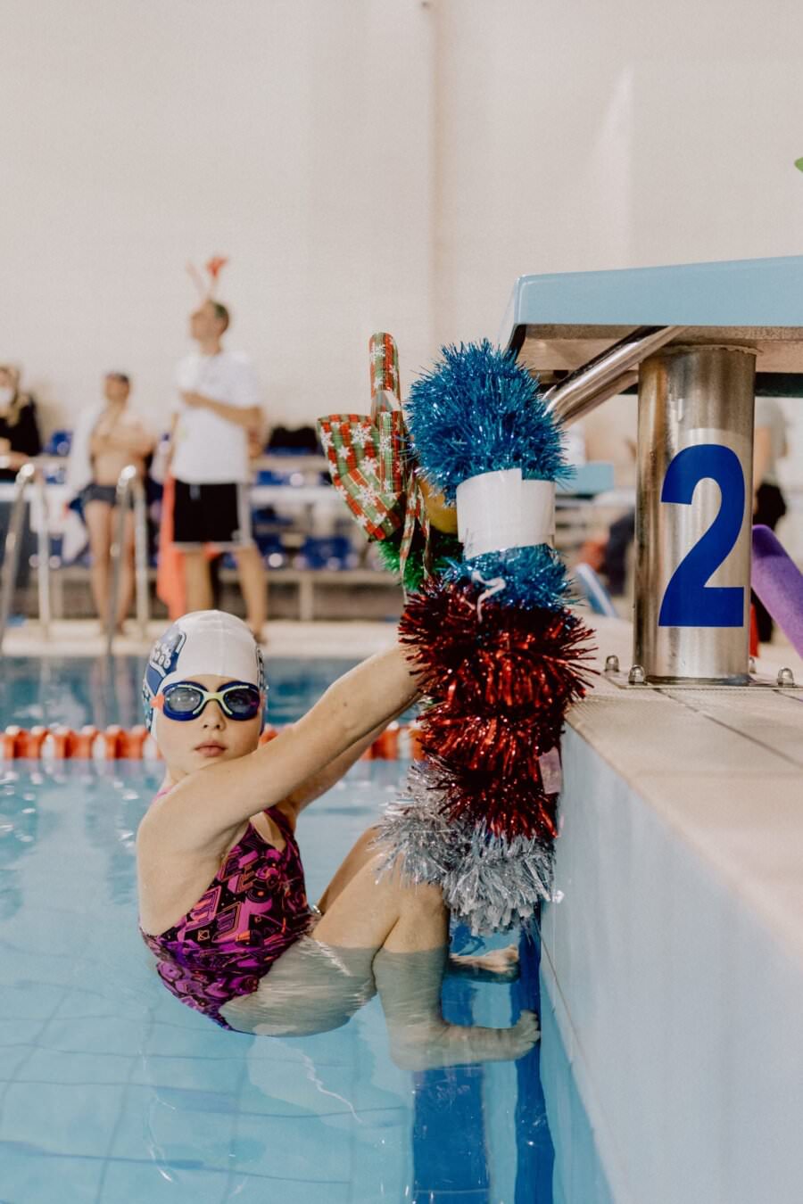 Młoda pływaczka w różowo-fioletowym kostiumie kąpielowym, czepku i okularach siedzi na brzegu basenu, trzymając się poręczy ozdobionej świątecznym świecidełkiem. W tle widoczne są znaczniki toru basenowego i inni pływacy. Teren basenu jest dobrze oświetlony, a jego jasna atmosfera została uchwycona przez Marcina Krokowskiego.