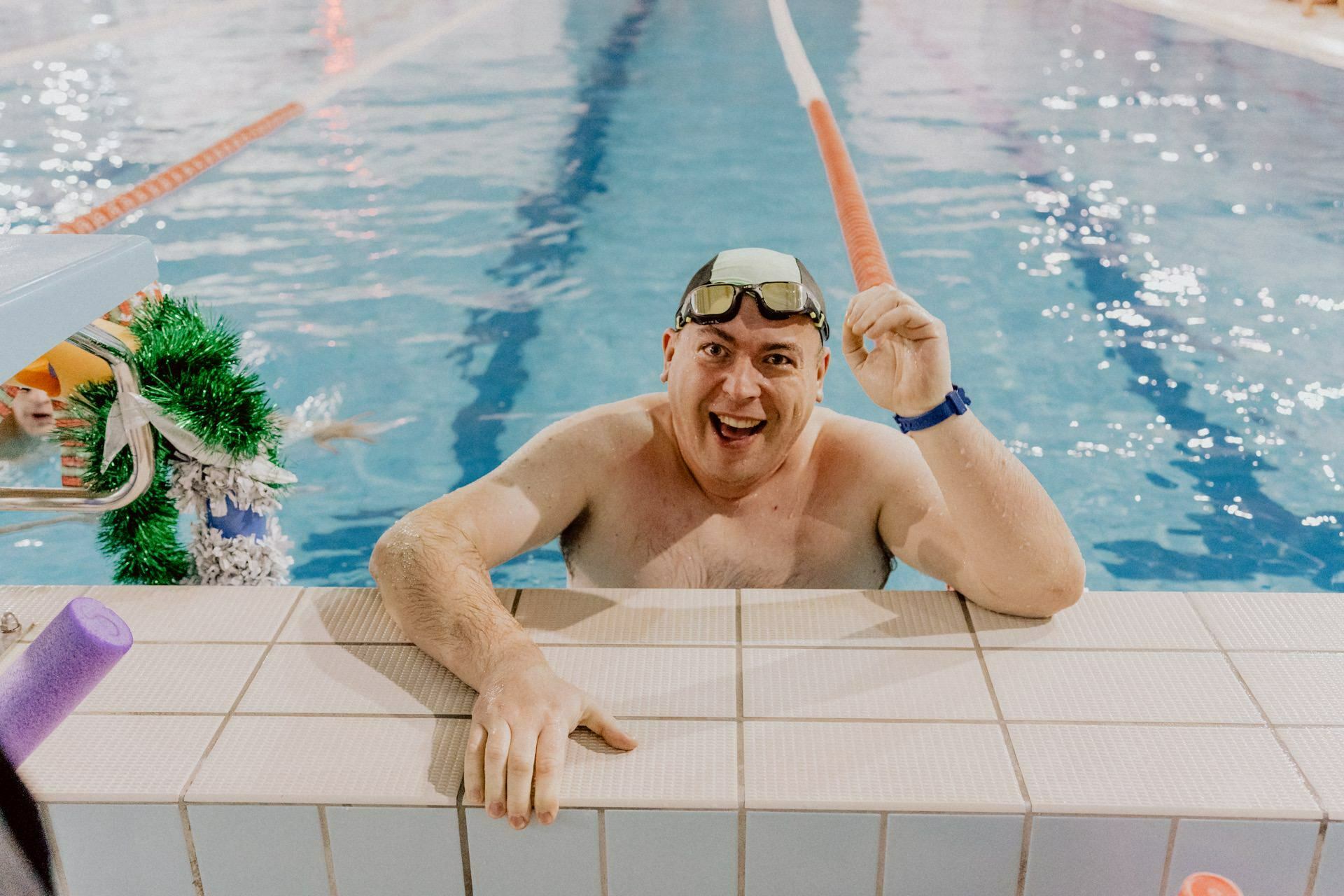 A smiling man wearing swimming goggles and a cap leans against the edge of the pool, holding a foam noodle. The pool has track splitters, and another swimmer is visible in the background. The scenery is reminiscent of an indoor pool, captured with the expertise of Marcin Krokowski.  