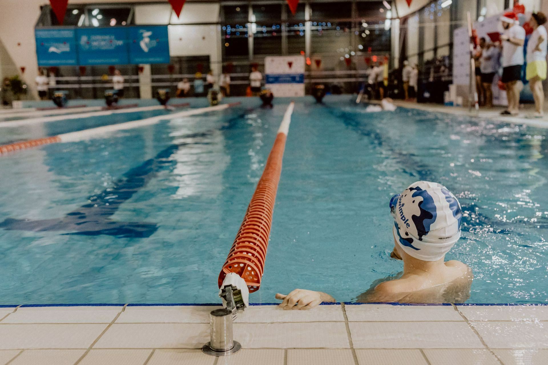 A swimmer wearing a white cap waits at the edge of the indoor pool, looking toward the lanes where other swimmers are competing. Spectators and activists stand on the side of the pool. The event, captured by well-known event photographer Marcin Krokowski, shows the red markings of the lane separating the pool.  