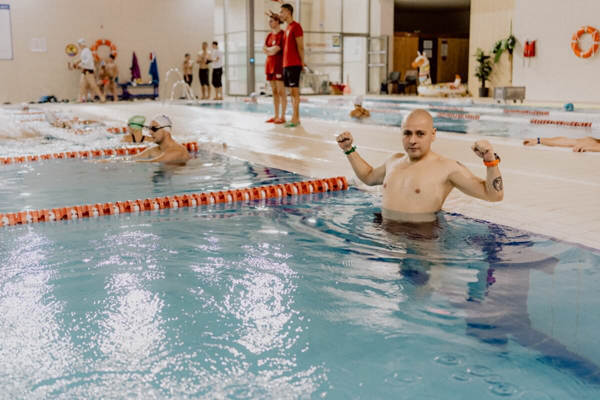 Osoba stoi w basenie blisko krawędzi, napina mięśnie ramion i uśmiecha się. W tle widać innych pływaków i inne osoby, niektórzy w wodzie, a niektórzy stoją przy basenie. Scena wydaje się być krytym obiektem pływackim, rejestrującym ożywioną fotorelację z wydarzenia.