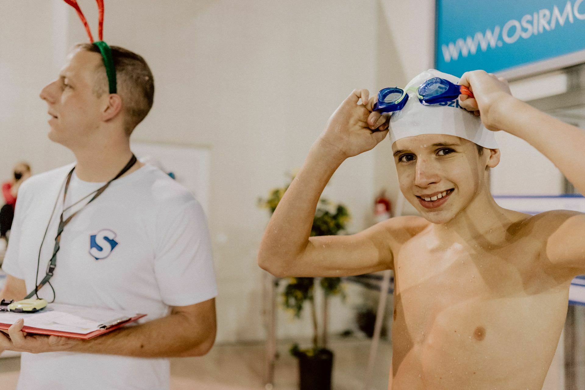 The swimmer smiles as he corrects his goggles, wearing a cap and standing next to a person in a white shirt with a logo, who is holding a notebook and wearing a Christmas headdress with horns. They are in a room, perhaps at a swimming competition or training, captured by Marcin Krokowski in an event photo. 