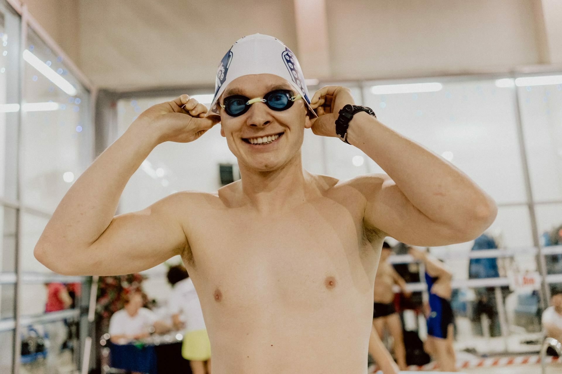 A shirtless person, wearing a white swim cap and blue goggles, smiles while standing in an indoor pool. They are correcting their goggles, while other swimmers and onlookers are in the background. This photo captures a vivid moment as part of a photo essay of the event.  