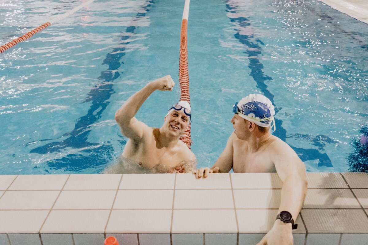 Dwóch pływaków w basenie stoi blisko krawędzi basenu, oddzielonych przegrodą toru. Pływak po lewej stronie podnosi prawą rękę w geście celebracji, podczas gdy pływak po prawej patrzy na niego, uśmiecha się, ma na sobie czepek i okulary pływackie. Idealne ujęcie dla każdego fotografa eventowego warszawy, aby pokazać żywe chwile.