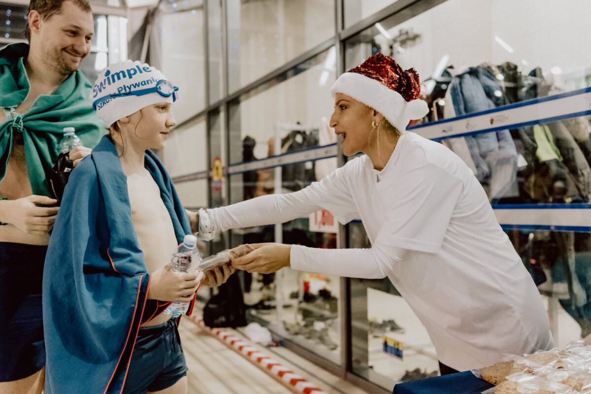 Kobieta ubrana w czapkę Mikołaja i biały t-shirt wręcza młodemu pływakowi butelkę wody i inne gadżety. Pływak okryty niebieskim ręcznikiem, w czepku i okularach pływackich przyjmuje z wdzięcznością. W pobliżu, w pomieszczeniu zamkniętym, stoi kolejna osoba w stroju kąpielowym, dzięki czemu jest to idealna fotorelacja z wydarzeń.
