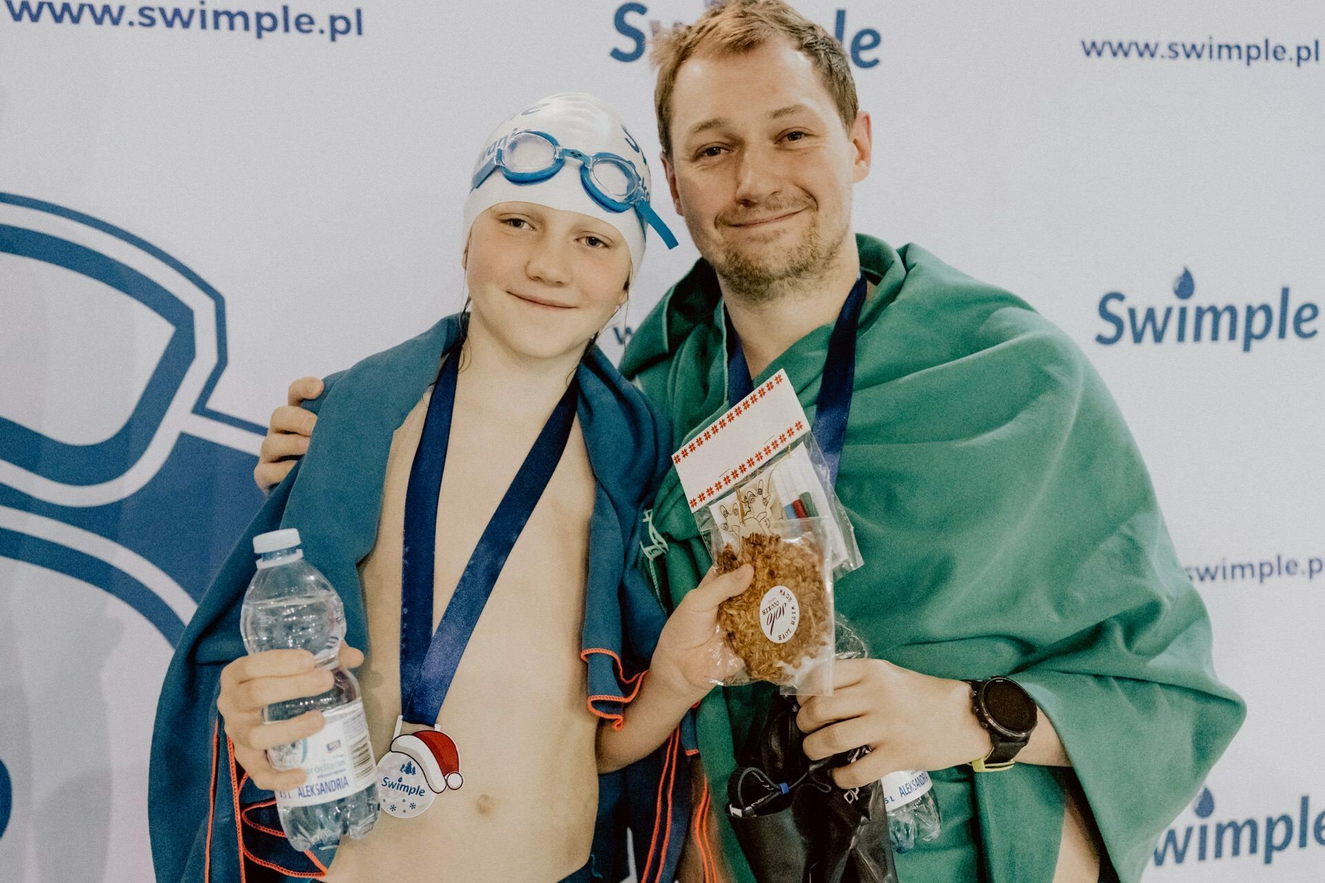 A smiling child wearing goggles and a swimming cap, wrapped in a towel, holds a bottle of water and a medal. Next to them stands an adult, also wrapped in a towel, holding a cookie and a small bag. They stand against a background of the "swimple.pl" logo by Marcin Krokowski, event photographer Warsaw.  