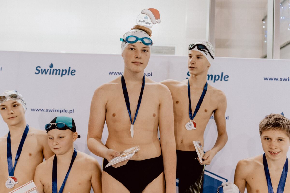 Grupa młodych pływaków ubranych w medale i czepki kąpielowe, w tym jeden z czapką Mikołaja na górze, stoi razem na tle „Swimple”. Trzymają drobne nagrody i noszą okulary do pływania, sprawiając wrażenie, jakby uczestniczyli w ceremonii wręczenia nagród lub wydarzeniu. Ta tętniąca życiem scena uchwyciła żywą fotorelację z eventu.