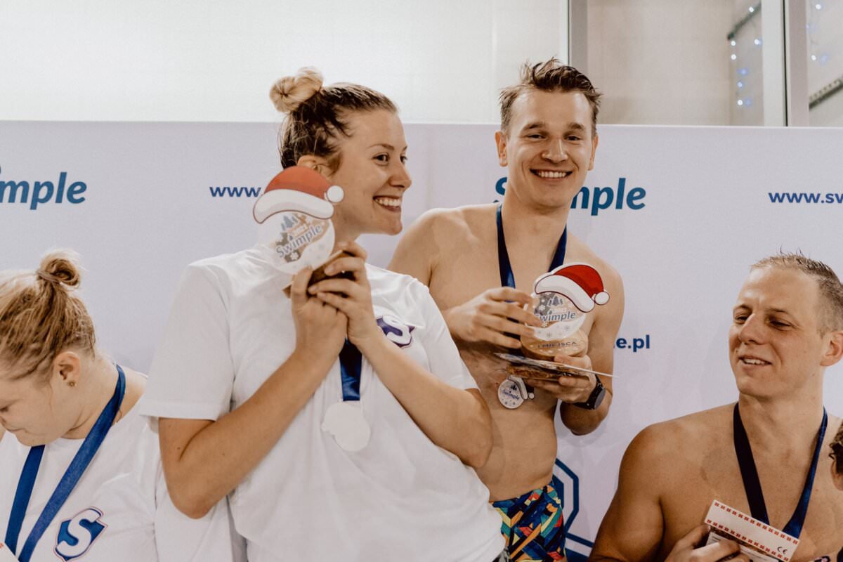 Czterech sportowców stoi na białym tle z tekstem „Próbka”, uśmiecha się i trzyma świąteczne nagrody z motywem Świętego Mikołaja. Oboje po lewej patrzą w dół, podczas gdy dwaj po prawej, kobieta i mężczyzna, stoją blisko siebie, promieniejąc i pokazując swoje medale – świetny przykład fotografii eventowej.
