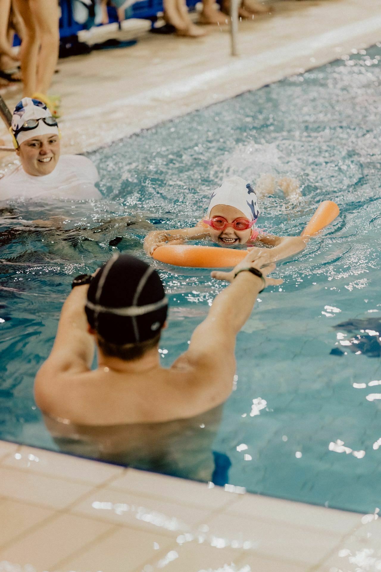 A young swimmer wearing red goggles and a white cap swims in a pool, using pool noodles, assisted by a coach wearing a black cap. Another swimmer, also wearing a white cap, watches from the background. The scene depicts an indoor pool, as documented by Marcin Krokowski's photo report of the events.  