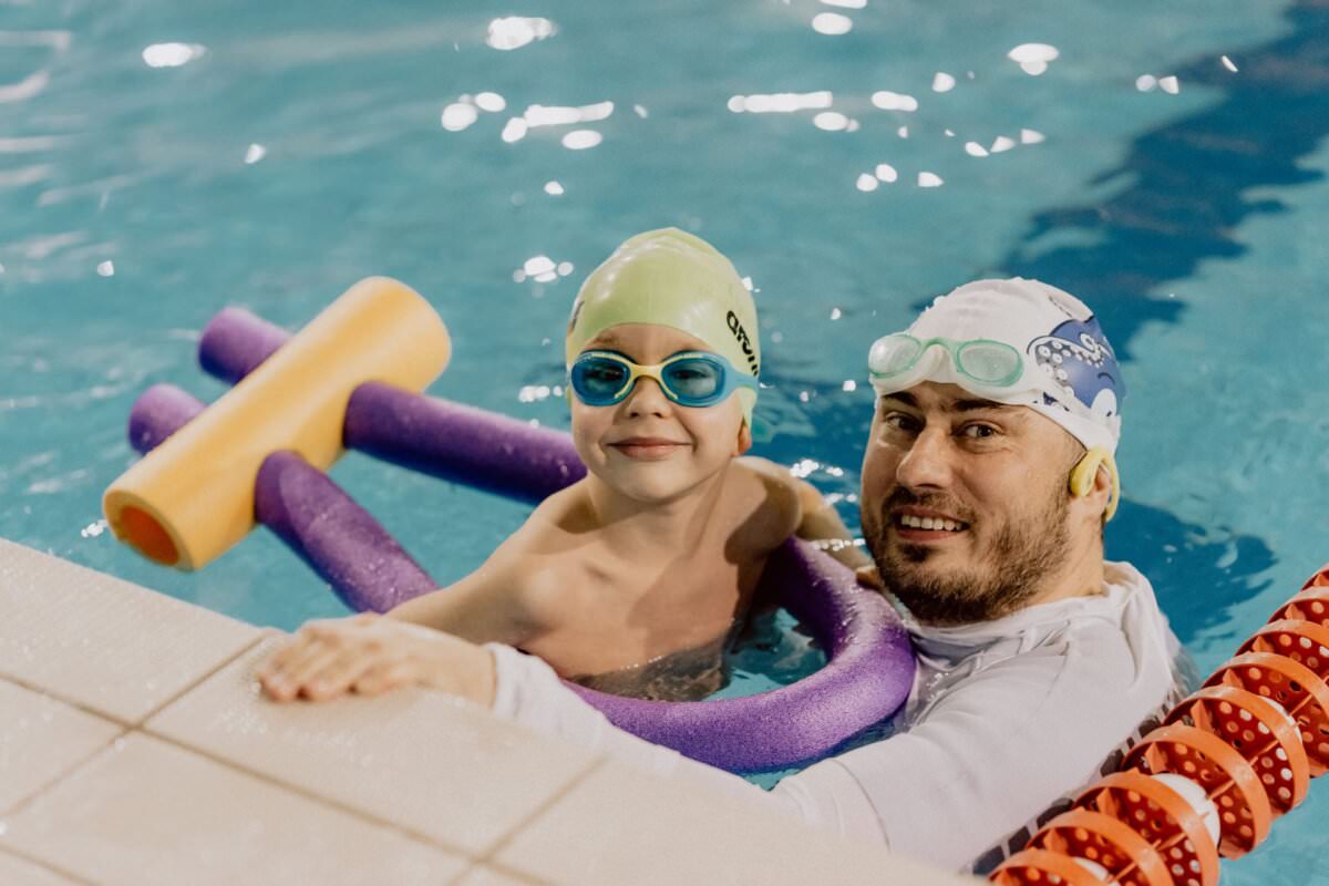 Dziecko w zielonym czepku i niebieskich okularach stoi w basenie, ściskając fioletowy makaron i uśmiechając się, obok mężczyzny w białym czepku i okularach, który również się uśmiecha. Znajdują się blisko krawędzi basenu, obok czerwonych i białych znaczników pasa. Fot. Marcin Krokowski.