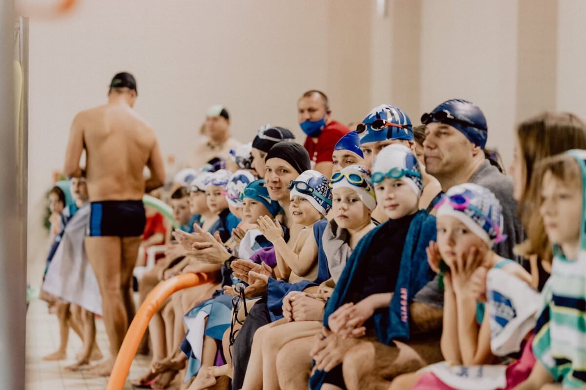 Grupa dzieci i dorosłych w czepkach i okularach pływackich siedzi na ławkach przy krytym basenie i ogląda wydarzenie pływackie. Niektóre są owinięte ręcznikami, inne mają sprzęt do pływania. Pływak w kostiumie kąpielowym stoi w pobliżu basenu, tyłem do kamery. Atmosferę obrazowo uchwycił Marcin Krokowski, utalentowany fotograf eventowy.