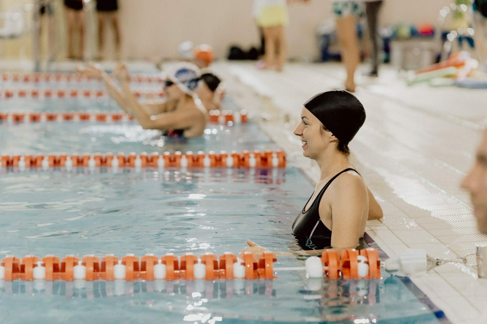 A person wearing a black cap and swimsuit stands in the pool, holding the edge. Other swimmers and pool equipment are visible in the background. The paths by the pool are separated by orange and white dividers, capturing the essence of event photography.  