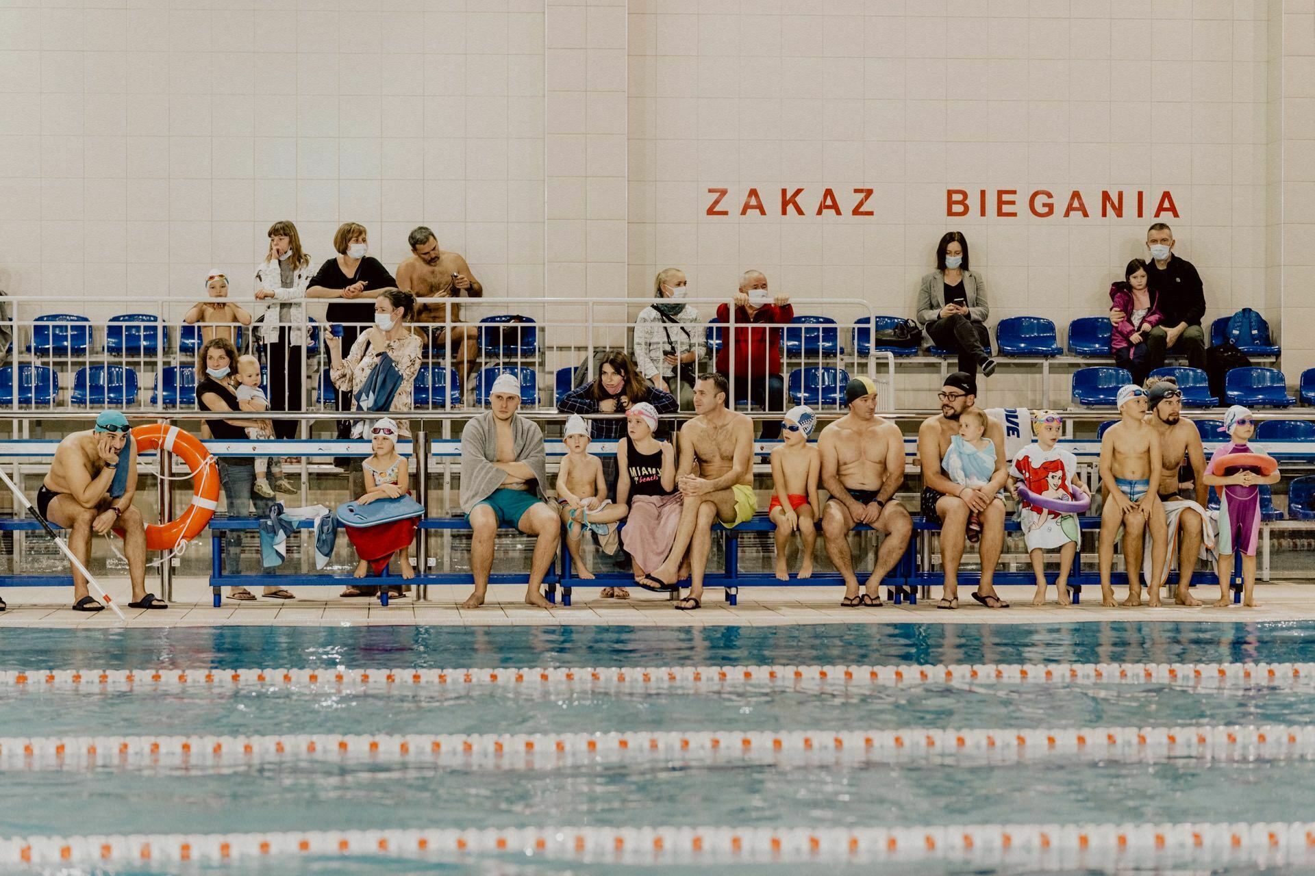 A group of swimmers sit on benches by the pool, wearing swimming caps and equipment. Behind them sit families and other spectators. A sign on the wall reads "No Running," which means "No Running" in Polish. The pool is in the foreground with the lane divisions, capturing a vivid photo report of the event.   